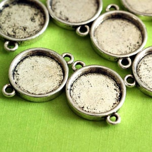 May include: Close-up of several silver-toned, round bezel settings with loop attachments. The settings have a textured, slightly aged appearance. The background is a bright green fabric, providing a contrasting backdrop for the jewellery components.