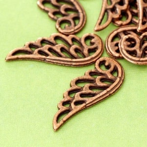 May include: Close-up of several bronze-coloured, openwork wing charms. The charms have a detailed, feather-like design and are arranged on a light green background. These decorative charms are ideal for jewellery making and craft projects.