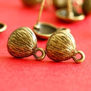 May include: Two antique brass-coloured, acorn-shaped beads with a textured surface and a small loop. The beads are on a red surface, with a blurred background showing other brass-coloured objects.