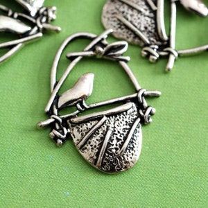 May include: Silver-coloured metal bird charm featuring a bird perched on a nest. The nest is textured and detailed, with a small bird sitting on a branch above it. The charm has a loop for attaching to jewellery or other crafts.