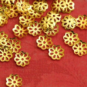 Könnte beinhalten: Eine Ansammlung kleiner, goldfarbener, blütenförmiger Metallkomponenten auf einer roten Oberfläche. Jede Komponente hat ein detailliertes, durchbrochenes Design mit blütenblattartigen Formen und einem zentralen Loch. Diese Komponenten werden wahrscheinlich in der Schmuckherstellung oder anderen Bastelprojekten verwendet.