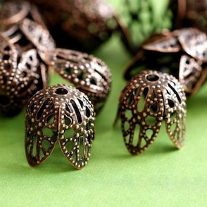 May include: Close-up of several bronze-colored filigree bead caps. The caps have an open, floral-like design with small, raised dots. The caps are on a light green surface, with some caps in the background out of focus.