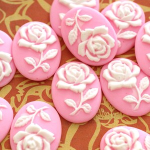 Puede incluir: Una colección de cabujones rosas ovalados con un diseño de rosa blanca en relieve. Las rosas tienen pétalos detallados y están sobre un fondo rosa liso y sólido. Los cabujones están dispuestos muy juntos.