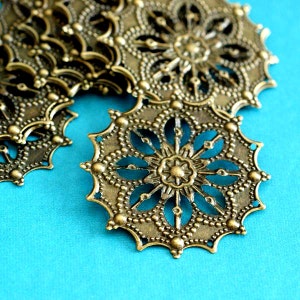 May include: Close-up of several antique brass filigree components with a floral design. The components are circular with intricate cut-out patterns and a central flower shape. The background is a bright teal color.