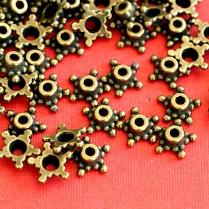 May include: Close-up of antique brass-coloured star-shaped metal beads. Each bead has a central hole and is surrounded by six points with small, decorative spheres. The beads are scattered on a red surface.