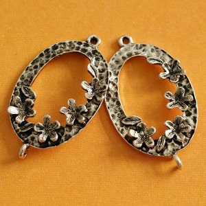 May include: Two silver oval pendants with a textured finish and floral embellishments. Each pendant has a loop at the top and a small hook at the bottom. The floral details are arranged around the oval's edge.