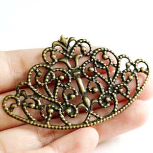 May include: An antique brass-colored filigree decorative piece. The ornate metalwork features a symmetrical design with swirling patterns and small circular accents. The piece has a curved shape, resembling a decorative drawer pull or furniture accent.
