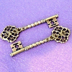 Puede incluir: Dos llaves de estilo antiguo en color bronce, con cabezas ornamentadas de filigrana y vástagos retorcidos. Las llaves están sobre una superficie púrpura. Las cabezas de las llaves tienen un diseño calado.