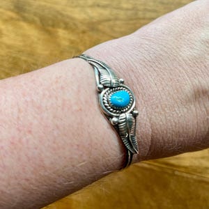 May include: A silver cuff bracelet with a central oval turquoise stone. The bracelet features leaf-like designs on either side of the stone and is worn on a wrist. The bracelet is made of a shiny metal.