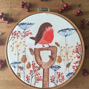 Peut inclure: Art de broderie en forme de cercle représentant un rouge-gorge perché sur une branche en bois brun. Le fond est un tissu blanc avec des baies rouges, des fleurs bleues et des accents dorés.