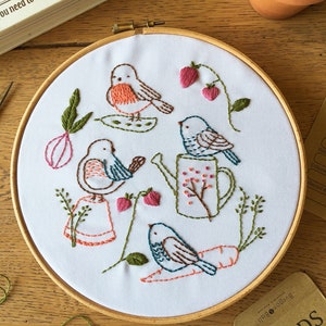 May include: Embroidered hoop art featuring several birds, strawberries, and a watering can. The design uses brown, orange, green, and pink thread on a white background. The hoop is set in a wooden frame. A book and seed packets are in the background.
