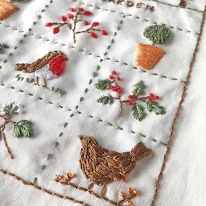 Puede incluir: Diseño bordado en tela blanca con un tema de jardín de invierno. El diseño incluye una casa, árboles, pájaros y bayas. El texto "Winter Garden" está bordado en hilo marrón.