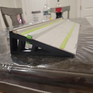 Może przedstawiać: Czarno-biała, kątowa prowadnica do obróbki drewna z zielonymi i srebrnymi akcentami. Prowadnica jest ustawiona na stole z przezroczystą plastikową osłoną. Prowadnica ma pomagać w precyzyjnym cięciu.