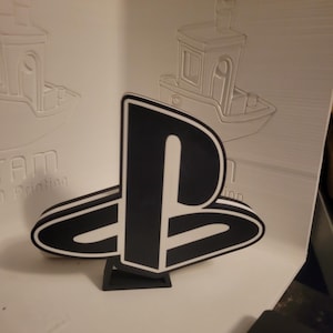 May include: A black and white PlayStation logo, 3D printed, stands on a small black base. The logo is in the shape of a stylized "P" and "S". In the background, there are embossed images of a boat.