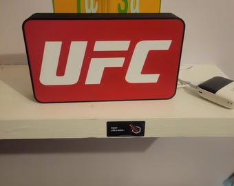 May include: A red and white sign with the word "UFC" in white letters. The sign is lit up and has a black border. There is a yellow sign with the text "Tam 3d" above the UFC sign. The signs are on a white shelf.