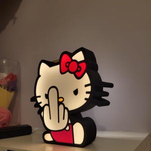 May include: A white Hello Kitty figurine with a red bow and black outline. The figurine is giving the middle finger with its right hand. The figurine is lit up from the inside.