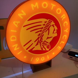 May include: A round, orange and yellow sign with the words "INDIAN MOTORCYCLE" and "1901" in white. The sign features a red and yellow image of a Native American wearing a headdress.