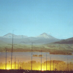 May include: A hazy, double-exposed photograph of a mountain range with power lines in the foreground. The mountains are in the distance and appear to be covered in snow. The power lines are in the foreground and are silhouetted against the sky. The image has a dreamy, ethereal quality.