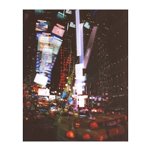 Könnte beinhalten: Ein verschwommenes Bild eines gelben Taxis, das nachts durch den Times Square in New York City fährt. Das Bild wurde aus dem Inneren des Taxis aufgenommen, mit Blick auf die Frontscheibe. Die hellen Lichter der Werbetafeln und Gebäude spiegeln sich in der Windschutzscheibe.