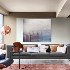 May include: A living room with a gray sofa, a coffee table, and a large framed print of a city skyline in the fog. The walls are painted in a light gray and a brown color. A copper globe pendant light hangs from the ceiling.
