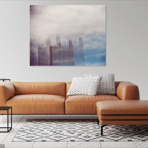 May include: A brown leather sectional sofa with a matching ottoman in a living room setting. The sofa has two white and gray patterned throw pillows. A large abstract painting of a city skyline in shades of blue, gray, and purple hangs on the wall behind the sofa.