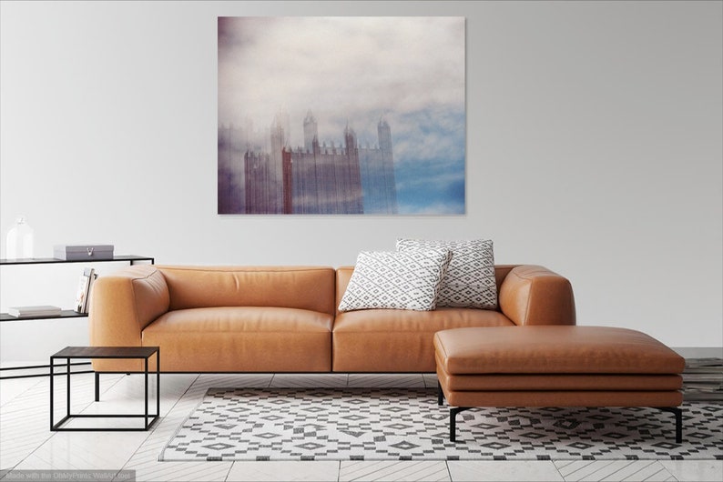 May include: A brown leather sectional sofa with a matching ottoman in a living room setting. The sofa has two white and gray patterned throw pillows. A large abstract painting of a city skyline in shades of blue, gray, and purple hangs on the wall behind the sofa.