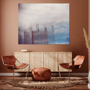 May include: A living room interior design with two brown leather chairs, a wooden cabinet with intricate carvings, a brown leather pouf, and a large canvas print of a city skyline in the fog.