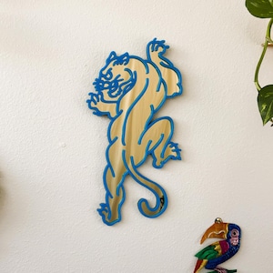 May include: A wooden tiger wall hanging with a light brown body and blue outline. The tiger is in a dynamic pose, with its front paws raised. The artwork is mounted on a white wall, alongside other decorative items.