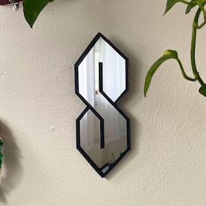 May include: A mirror in the shape of the stylized "S" symbol, with a black frame, mounted on a cream wall. The mirror reflects a window and is surrounded by other wall decorations, including plants and a wreath.