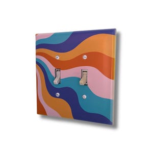 Può includere: Una placca per doppio interruttore della luce con un motivo ondulato multicolore nei toni dell'arancione, rosa, verde acqua e blu scuro. La copertura rettangolare include due interruttori a levetta verticali color beige chiaro e quattro viti argentate.