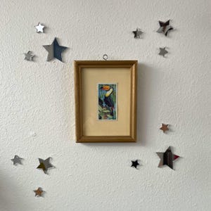 May include: A rectangular gold-toned picture frame containing a vertical print of a toucan perched on a branch amidst green foliage. The frame is surrounded by several small, reflective, silver-colored star-shaped wall decals of varying sizes.