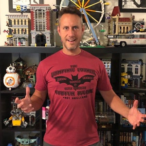 Puede incluir: Un hombre con una camiseta roja con un estampado gráfico negro que dice "The Vampiric Council" con un logotipo de murciélago y el texto "Staten Island - Not Guillermo".