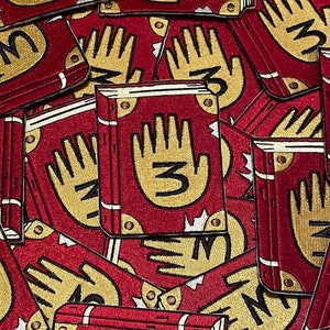 Könnte beinhalten: Ein Stapel roter gestickter Patches mit einer goldenen Hand mit der Nummer 3 darauf. Die Patches haben alle das gleiche Design und sind übereinander gestapelt.