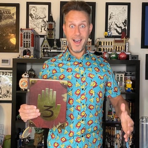 May include: A person wearing a blue shirt with a yellow triangle pattern and a red book with a hand and the number 3 on the cover. The book is titled "Journal 3".
