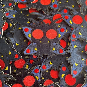 May include: A close-up of a pile of embroidered patches featuring a cartoon monster with red eyes, blue wings, and yellow accents. The monster is set against a dark blue background.
