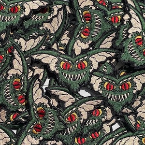 May include: Embroidered patches featuring a green creature with large red eyes, sharp teeth, and bat-like wings. The patches have a detailed design with tan and green stitching, creating a textured appearance. The patches are likely for clothing or accessories.