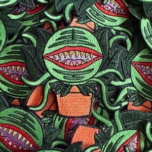 Puede incluir: Parches bordados con una planta verde de estilo caricatura con una gran boca dentada. Los parches tienen macetas naranjas y hojas de color verde oscuro. La boca tiene labios rojos y dientes blancos. Los parches están densamente empaquetados.