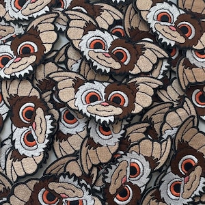 May include: A close-up of a pile of brown and white embroidered patches featuring a cartoon creature with large eyes and bat-like ears. The patches are all identical and are arranged in a random pattern.