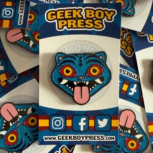 Könnte beinhalten: Ein blauer Glitzer-Emaille-Pin mit einer katzenartigen Kreatur mit roten und gelben Augen, einer rosa Zunge und scharfen Zähnen. Der Pin befindet sich auf einer Trägerkarte mit dem Text "GEEK BOY PRESS" und Social-Media-Symbolen. Die Website www.geekboypress.com ist ebenfalls sichtbar.