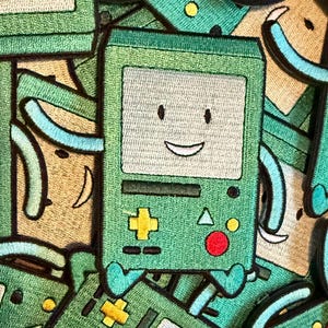 May include: Green embroidered patch featuring a smiling, square-shaped character with a light beige face, a yellow plus symbol, and a red circle. The character has small blue feet and arms. The background is filled with similar patches.