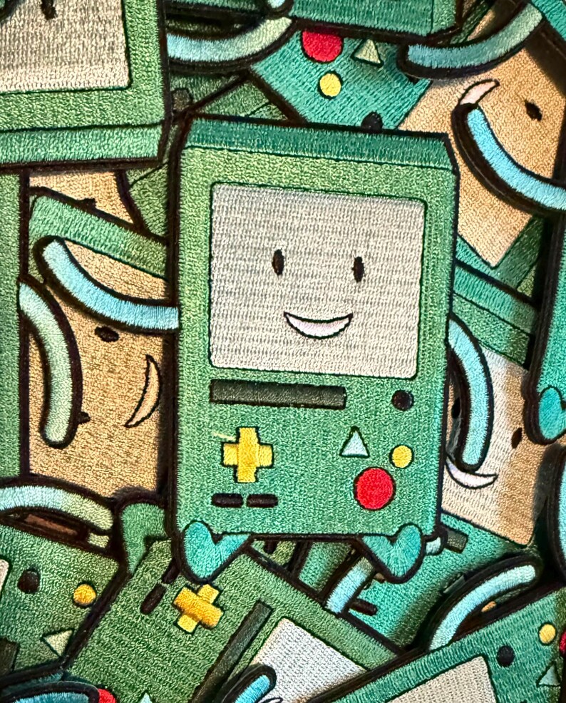 May include: Green embroidered patch featuring a smiling, square-shaped character with a light beige face, a yellow plus symbol, and a red circle. The character has small blue feet and arms. The background is filled with similar patches.