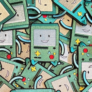 May include: A collection of green and tan embroidered patches featuring a smiling, retro-style video game console character. The patches have a light blue screen, red and yellow accents, and a happy face.