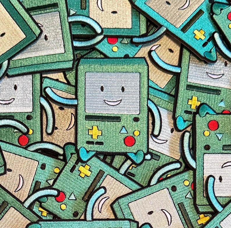 May include: A collection of green and tan embroidered patches featuring a smiling, retro-style video game console character. The patches have a light blue screen, red and yellow accents, and a happy face.