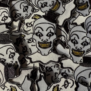 Può includere: Una collezione di toppe ricamate raffiguranti una faccia di clown sorridente, completa di cappello a cilindro nero e colletto. Il volto del clown è bianco con contorni neri e denti gialli. Le toppe sono fittamente raggruppate.