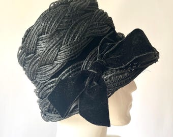 May include: A black vintage hat with a woven design and a velvet bow. The hat has a rounded crown and a small brim. The bow is positioned on the side of the hat. The hat is displayed on a mannequin head.