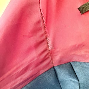 May include: Close-up of a garment featuring a deep red fabric with a subtle sheen, stitched to a section of navy blue material. The stitching is visible, and a small black ribbon is present.
