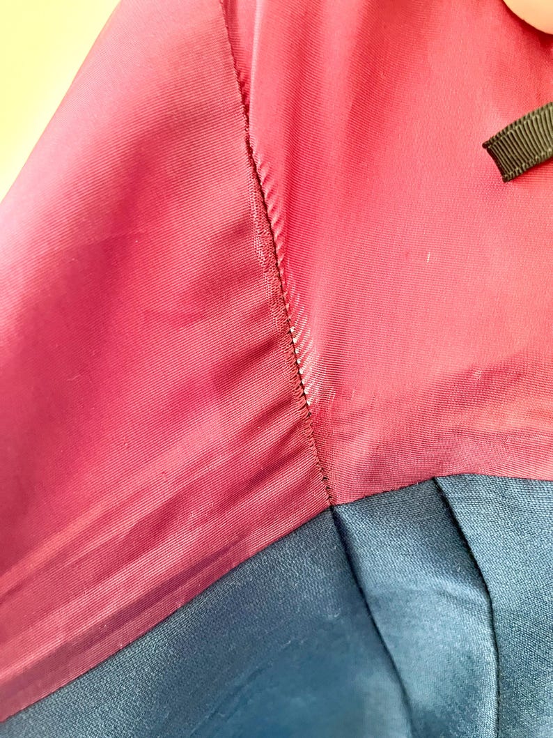 May include: Close-up of a garment featuring a deep red fabric with a subtle sheen, stitched to a section of navy blue material. The stitching is visible, and a small black ribbon is present.