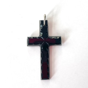 May include: A black cross pendant with a textured surface and a silver-colored bail. The cross has a dark red interior visible through the textured design. The pendant is a religious symbol.