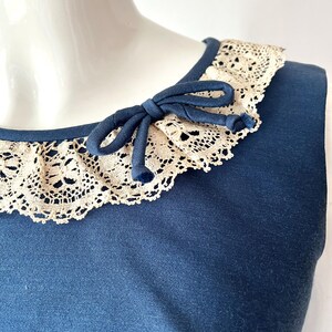 May include: A sleeveless navy blue top with a cream-colored lace collar. A navy blue bow is tied at the neckline. The lace collar adds a vintage touch to the garment.