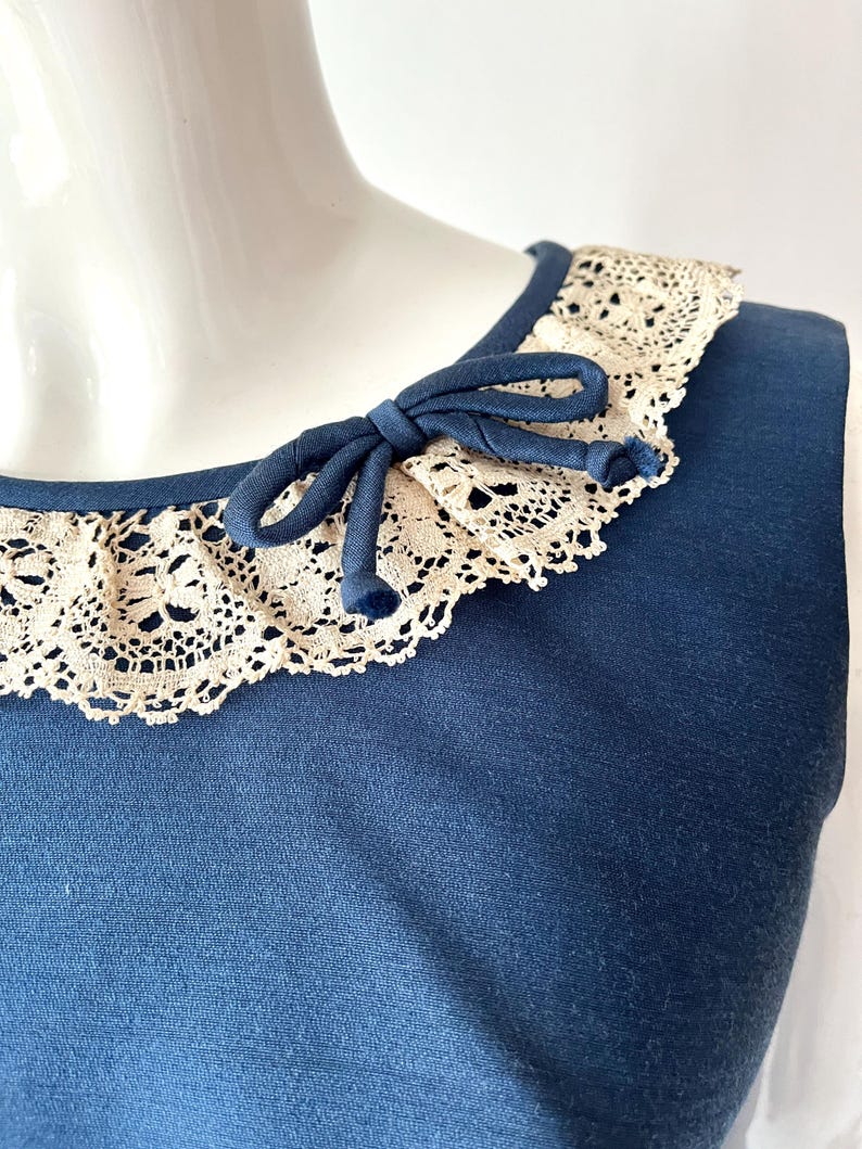 May include: A sleeveless navy blue top with a cream-colored lace collar. A navy blue bow is tied at the neckline. The lace collar adds a vintage touch to the garment.